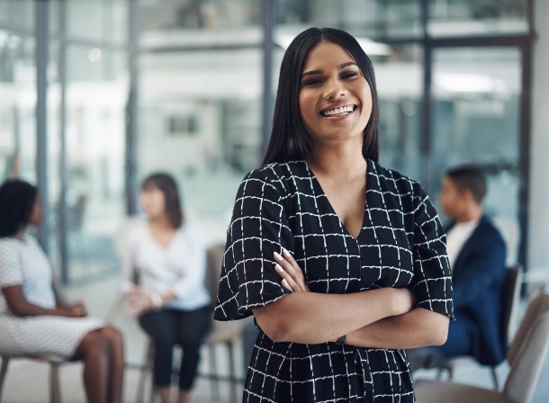 Female Brown Employee smiling in an office setting with other employees out of focus behind her.
